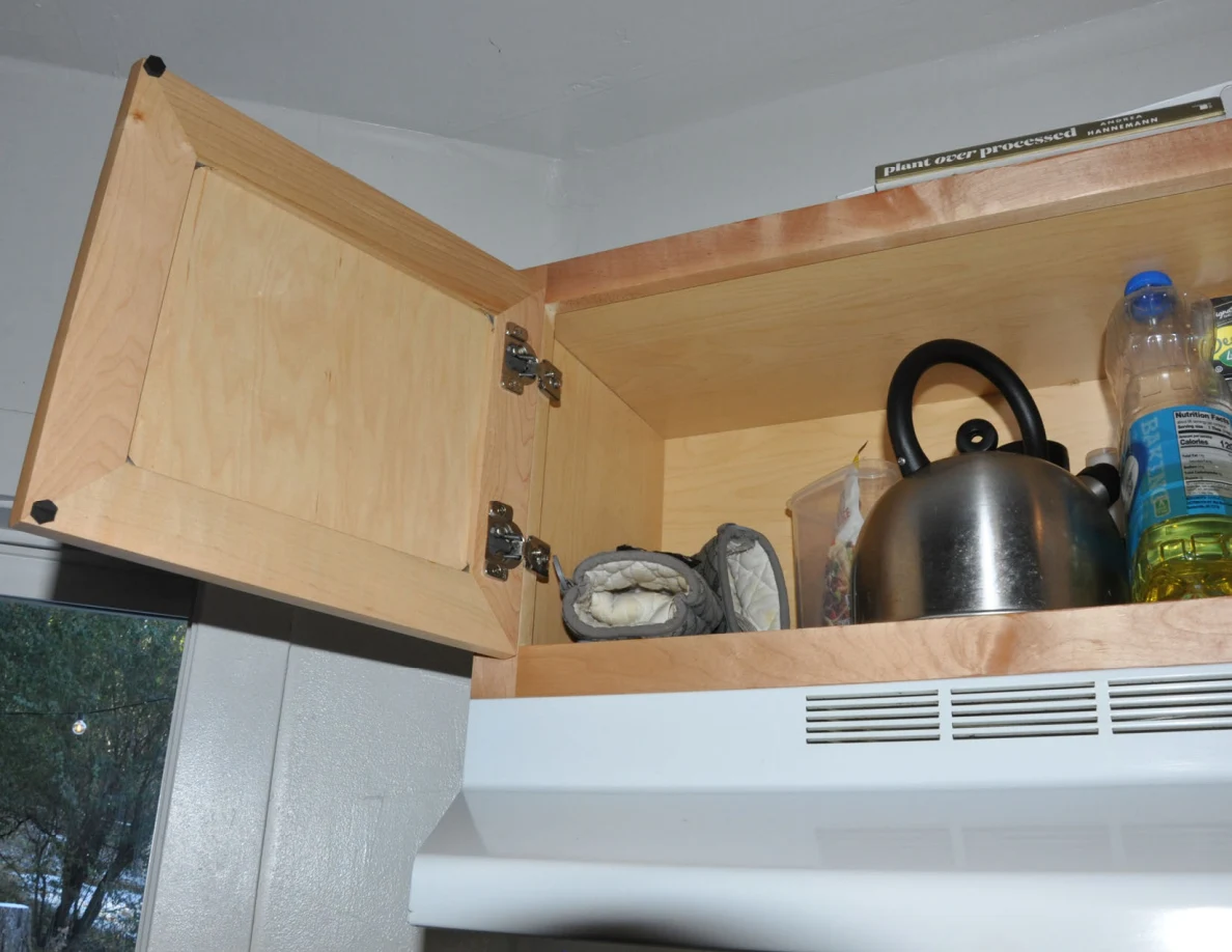 Open cabinet above range hood containing a metal kettle, bottle of oil and cloths.