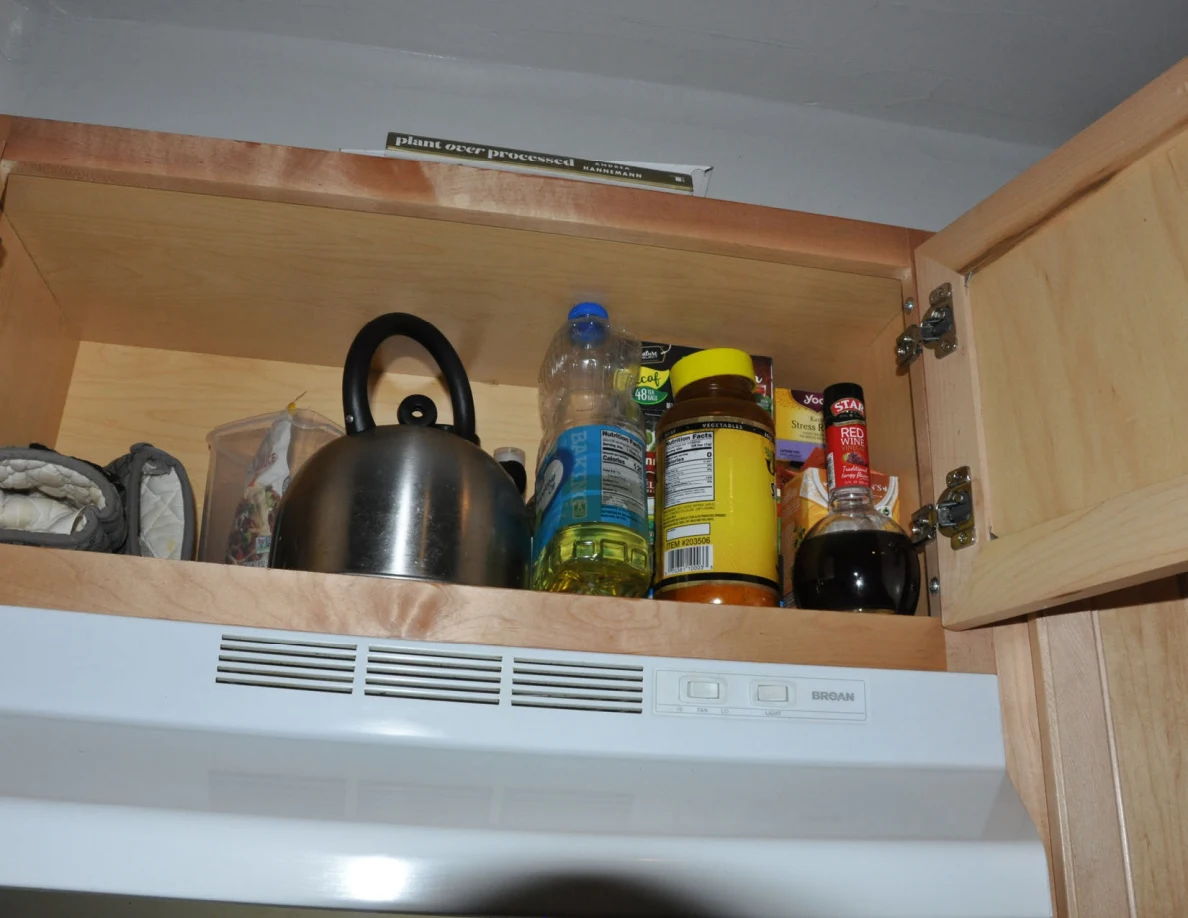 Open cabinet above range hood with metal kettle, oil bottle, jars and condiments.