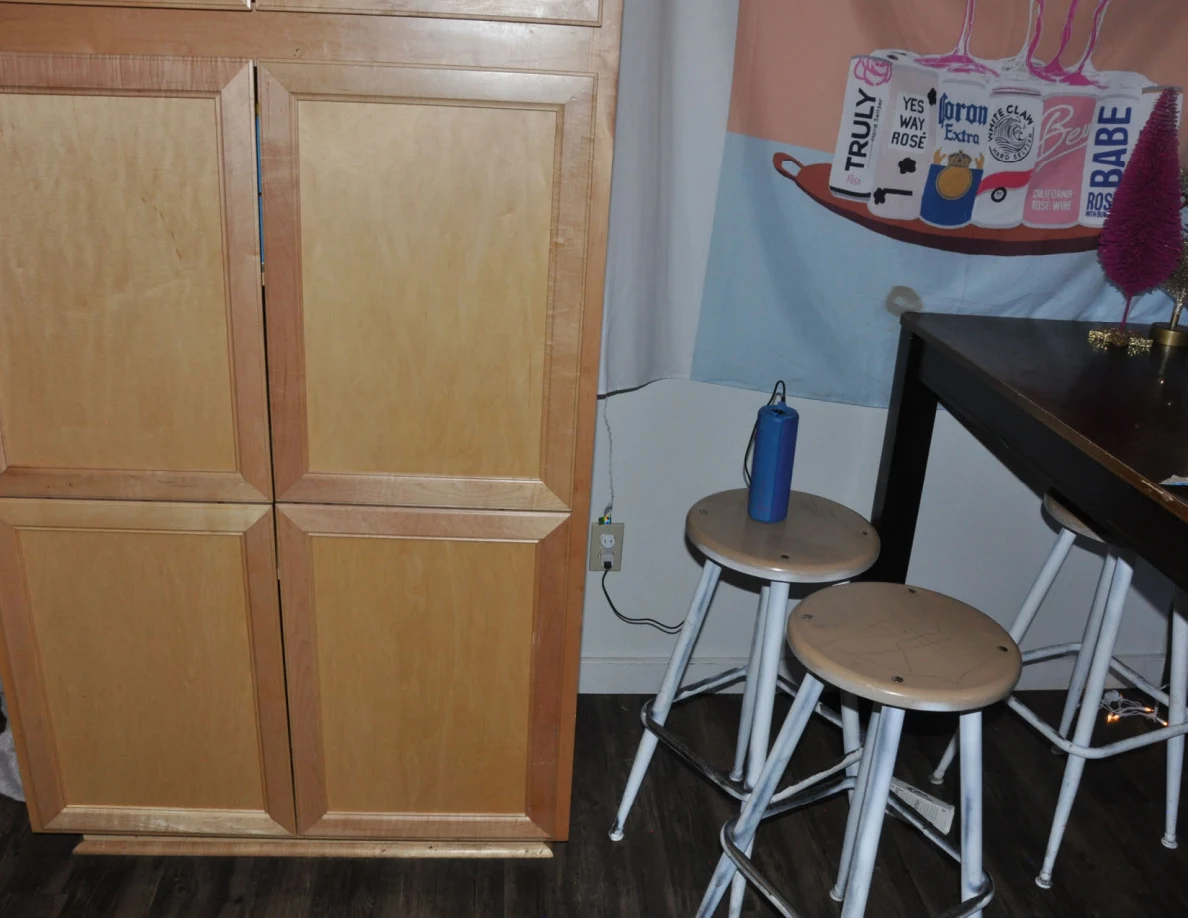 Closed wooden pantry cabinet beside stools, blue speaker and kitchen table.