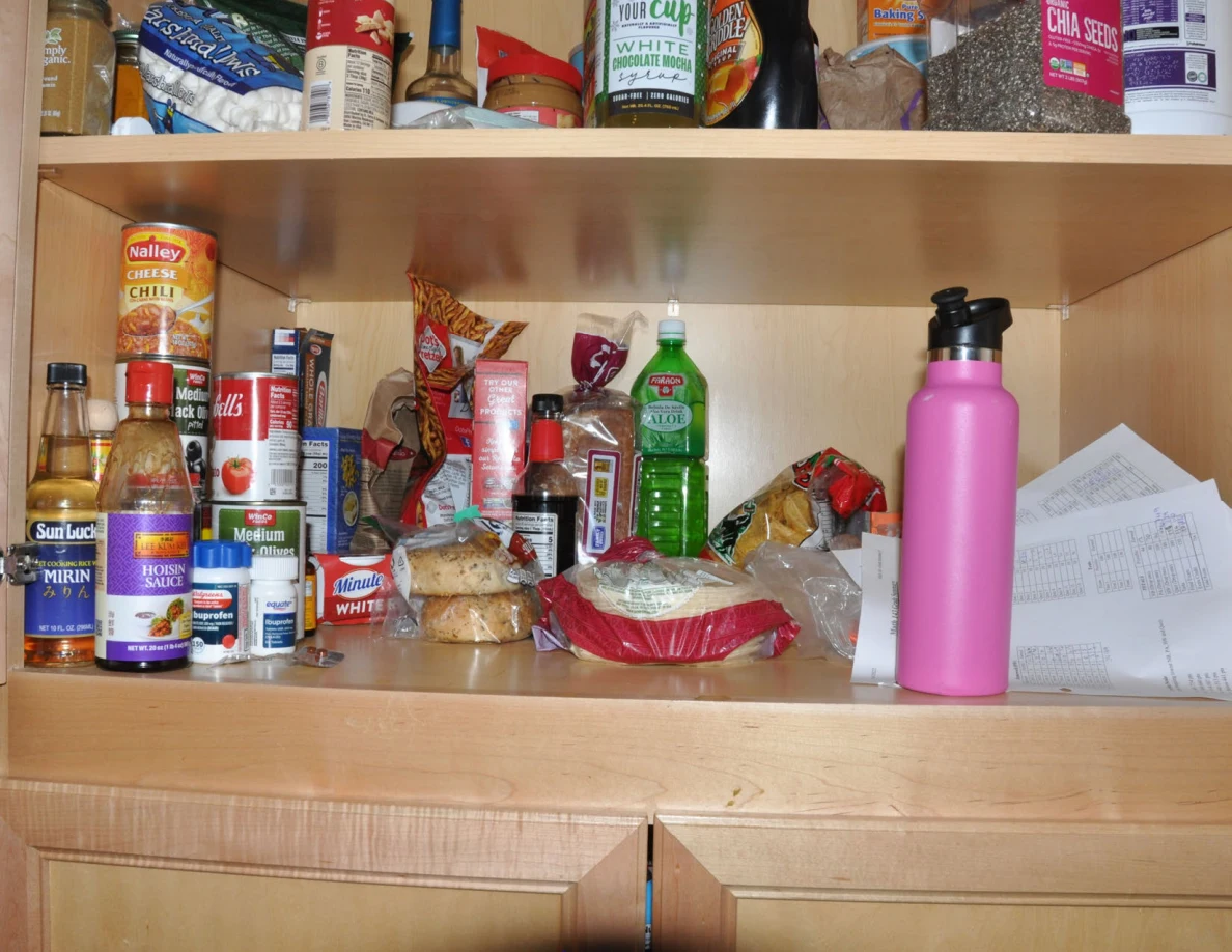 Pantry shelf with sauces, canned food, bread packages, papers and a pink bottle.