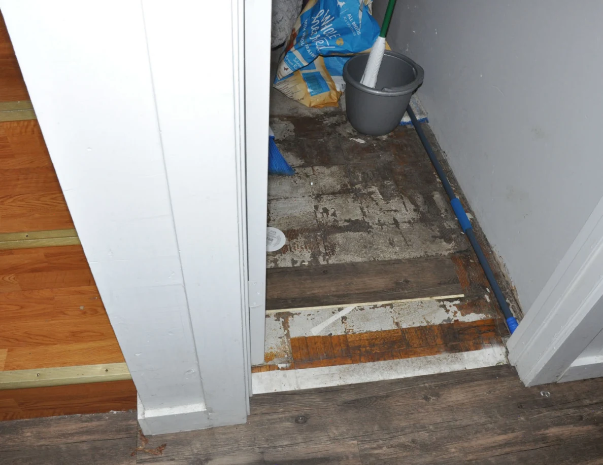 Open storage closet threshold with a mop bucket and bags on a worn floor.
