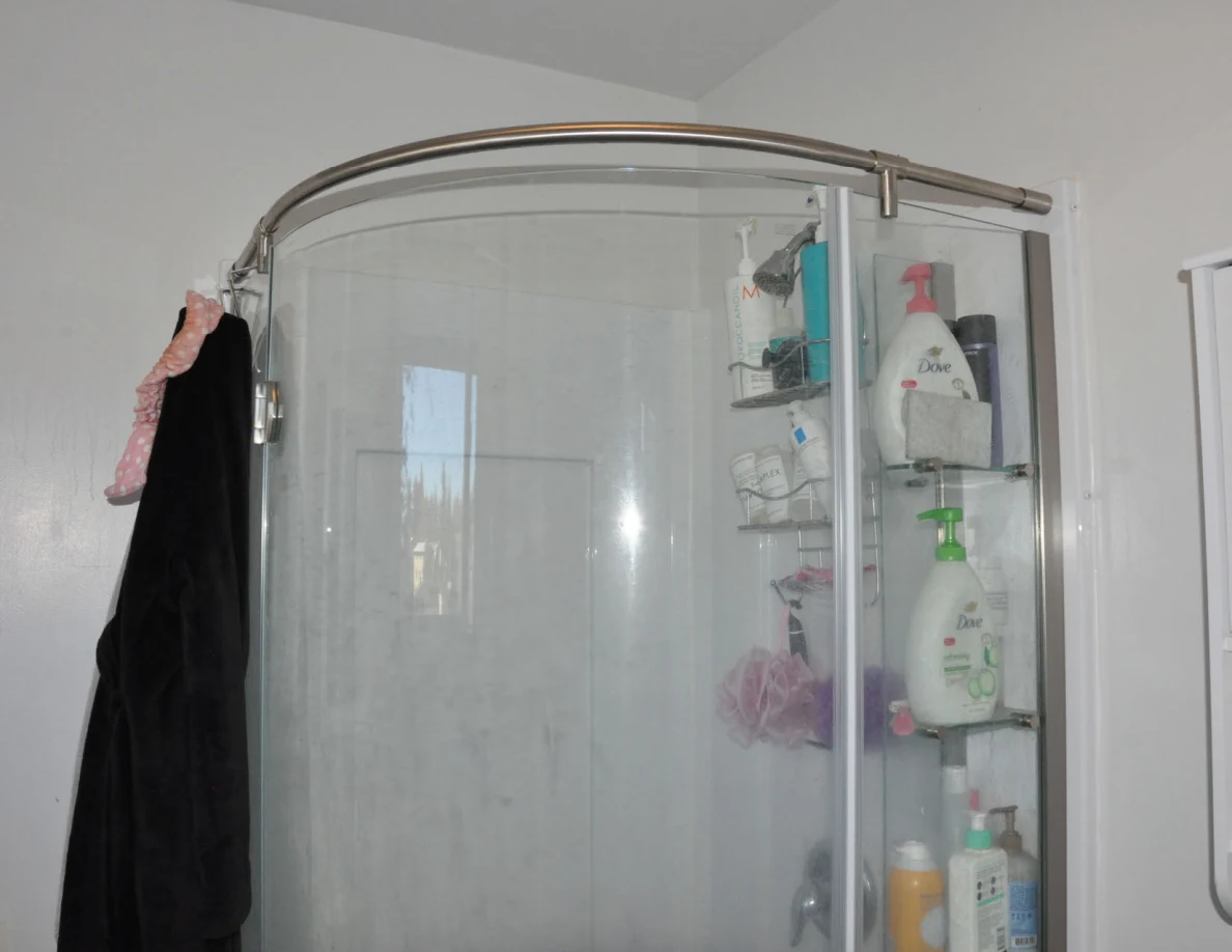 Curved glass shower enclosure with shelves of toiletries and a dark robe hanging beside it.