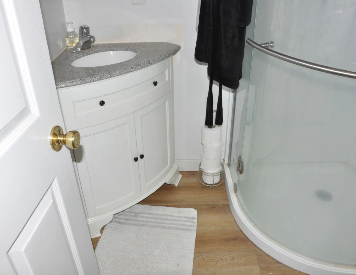 Curved white bathroom vanity and glass shower beside a bath mat.