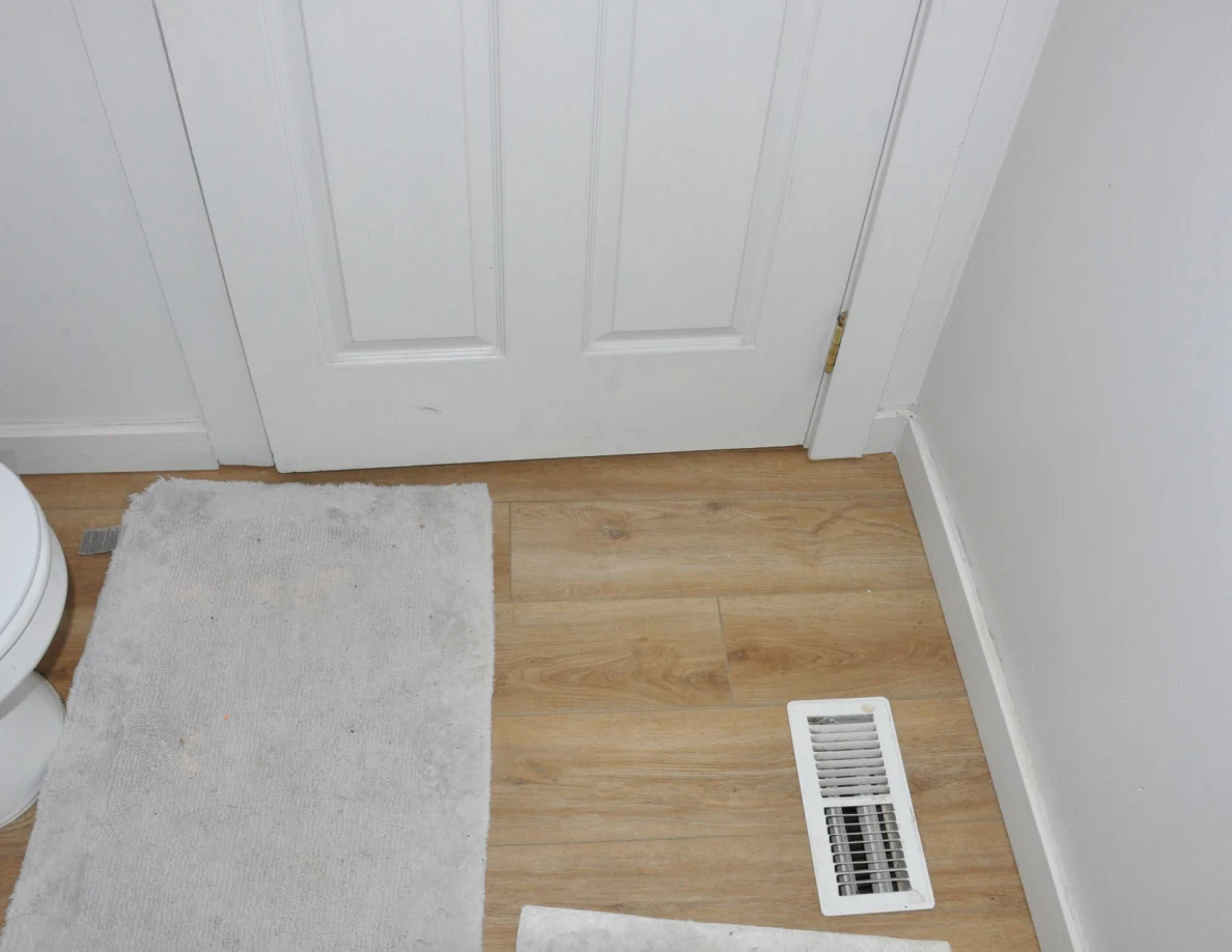 Lower white bathroom door beside a pale bath mat and floor vent.