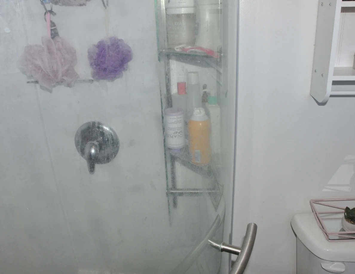 Glass shower door with faucet, bath poufs and a corner shelf of toiletries.