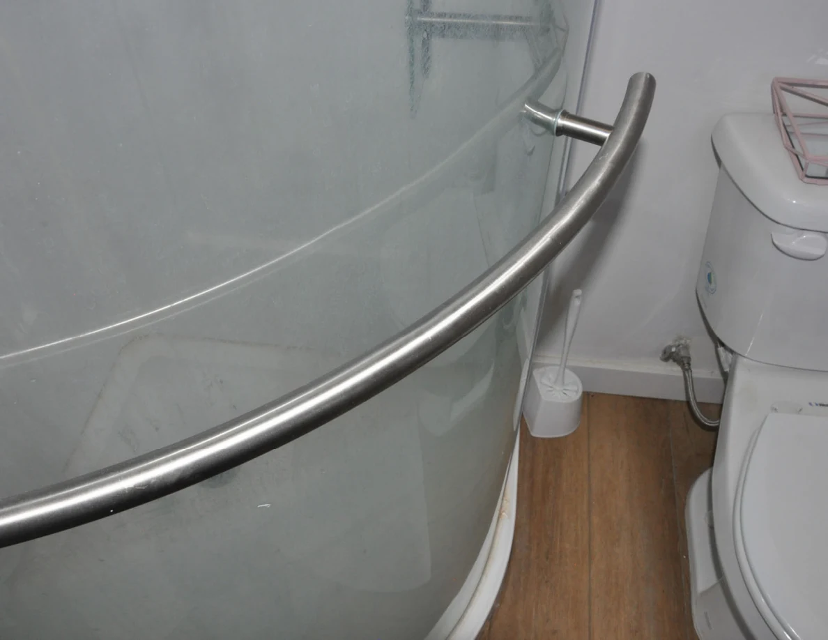 Close view of curved metal shower-door handle beside a toilet.