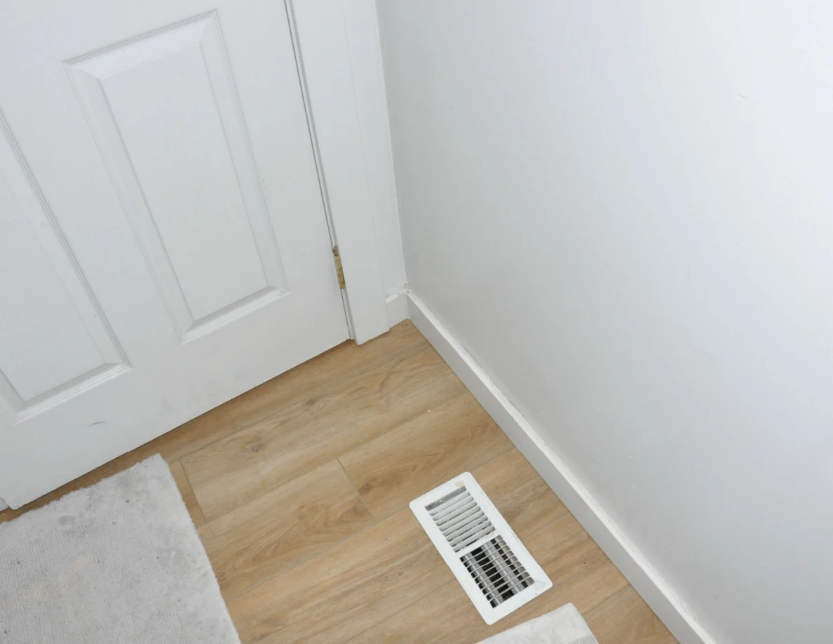 Floor vent beside a bathroom door and pale bath mat on wood-look flooring.