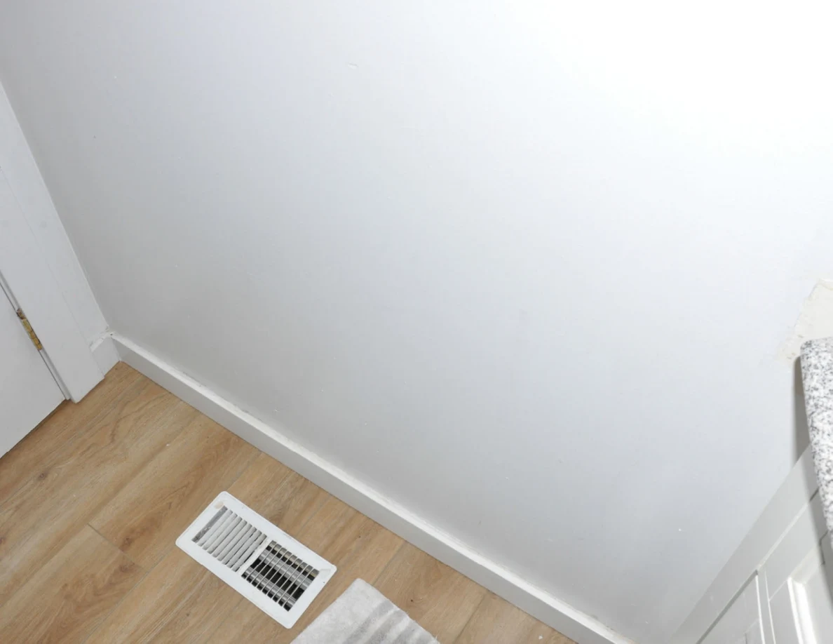 Bathroom floor vent beside white wall, wood-look flooring and the edge of a bath mat.