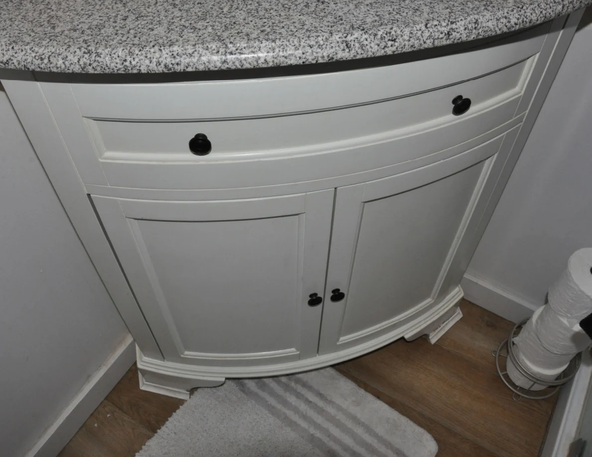 Curved white vanity cabinet with black knobs beneath a gray-speckled counter, with a mat below.