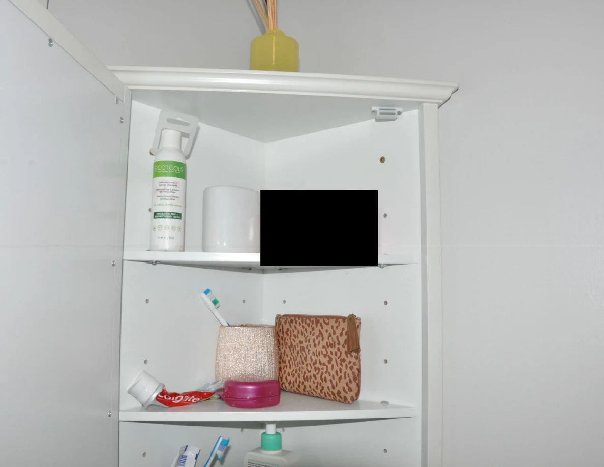 Open medicine cabinet containing bottles, a cup, toothbrush, toothpaste and patterned pouch, with a black redaction.