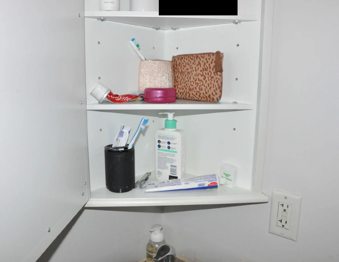 Lower shelves of an open medicine cabinet with toothbrushes, toothpaste, lotion bottle, cups and pouch, with a black redaction above.