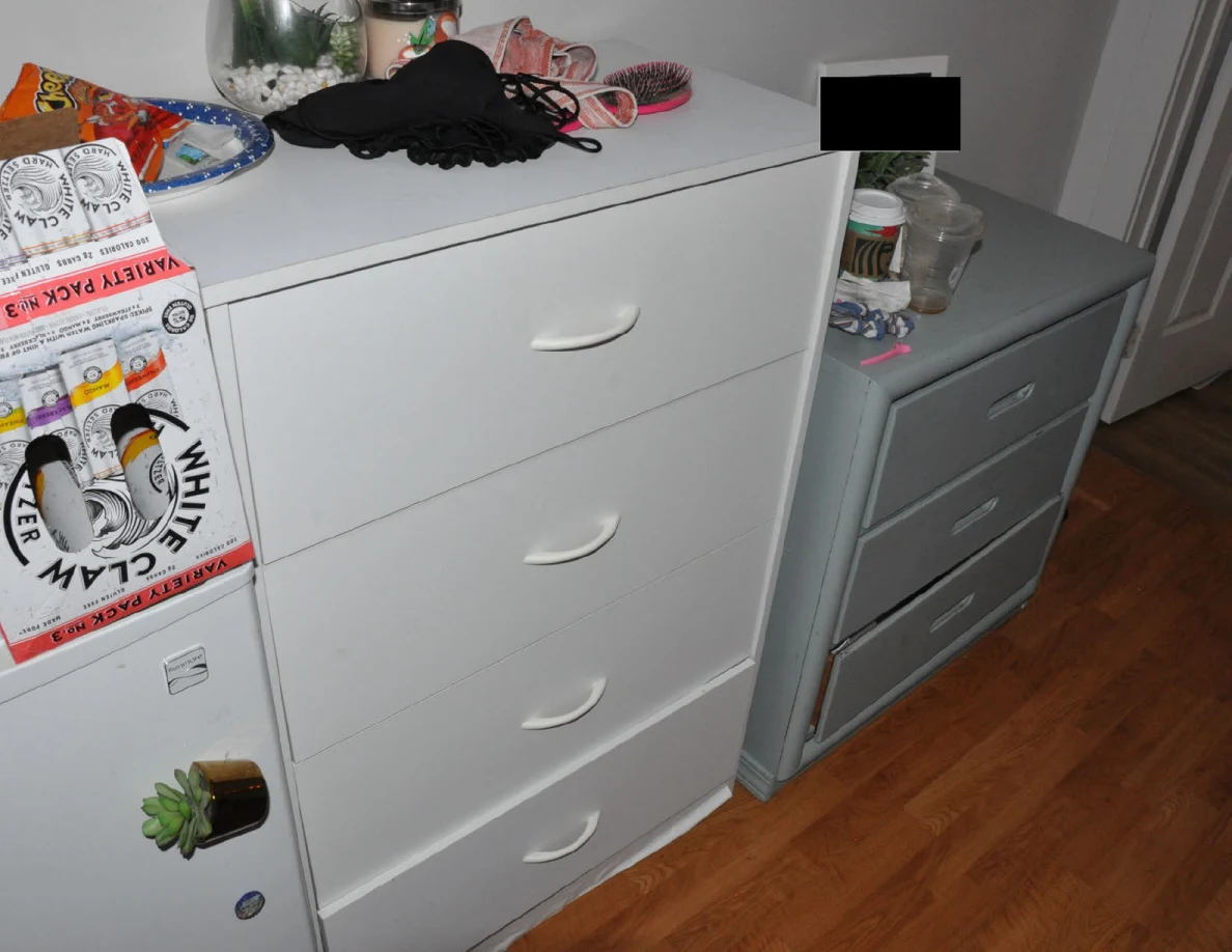 Closed white and gray dresser drawers with a hairbrush and personal items on top, a drink carton at left and a redacted frame.