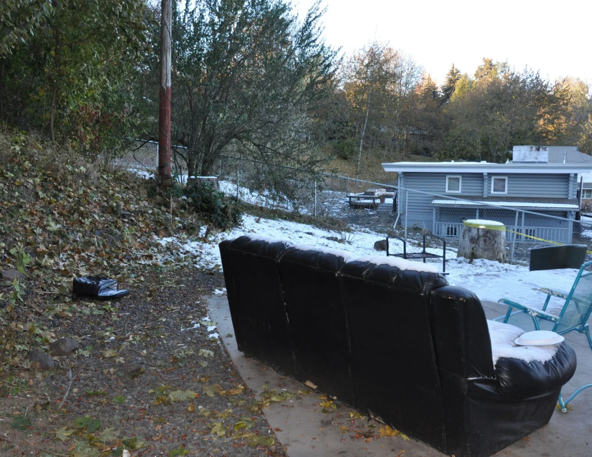 Snow-dusted black sofa outdoors beside a patio, with trees and a neighboring house beyond.
