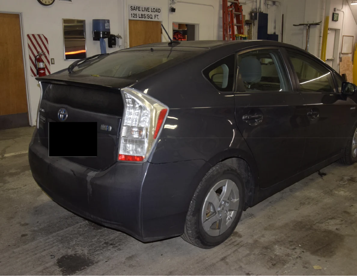 Opposite rear corner of a dark blue Toyota Prius, showing rear lights and a blacked-out plate.