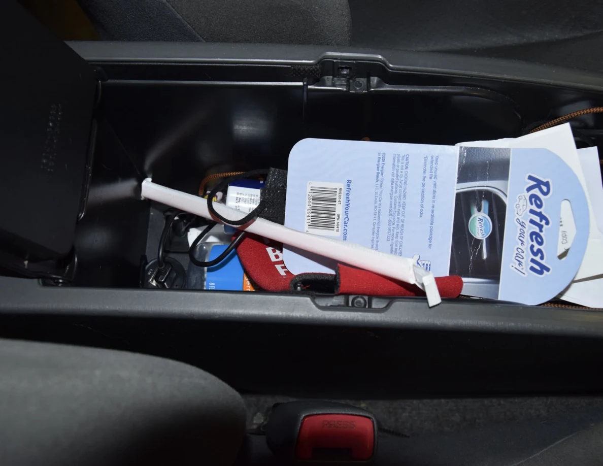 Open center-console compartment with air-freshener packaging, a white tube and cables.