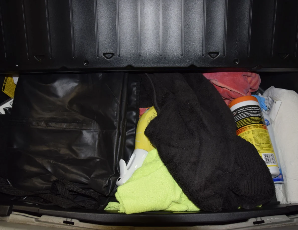 Second view of under-floor hatch storage with a black bag, towels and cleaning-product can.