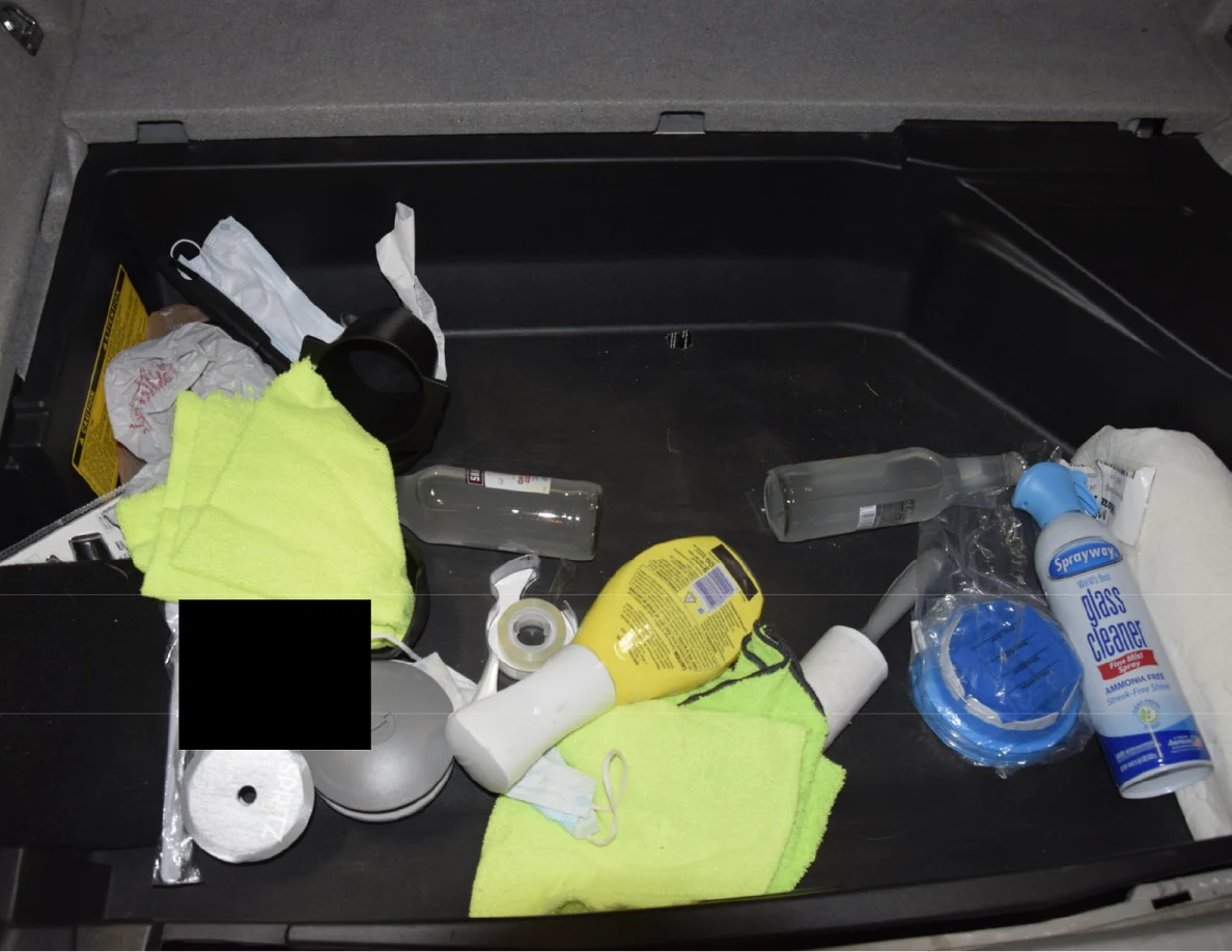 Hatch storage containing yellow cloths, spray bottles, glass cleaner, plastic bottles and a black redaction.