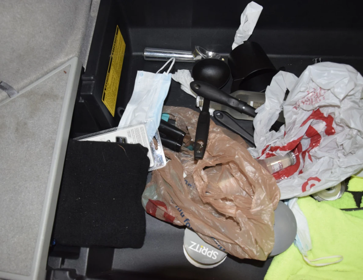 Under-floor storage with plastic shopping bags, black utensils, disposable face mask and black knit fabric.