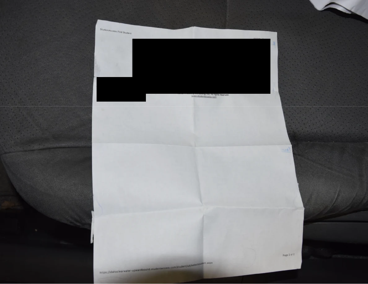 Folded printed paper on a gray car seat with black redactions across its upper portion.