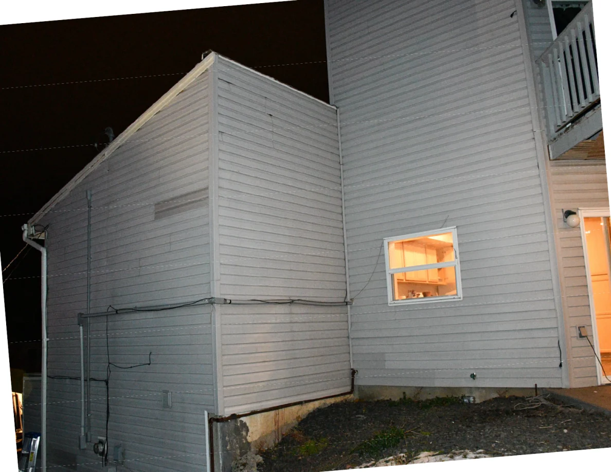 Exterior corner of a gray-sided house at night, with a lit window and upper balcony edge.
