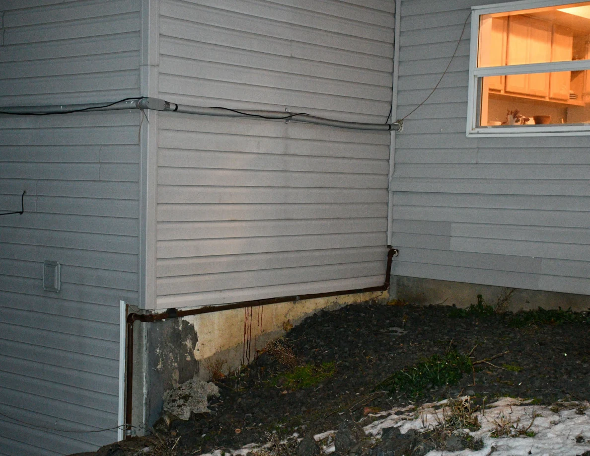 Gray exterior siding, pipes and concrete foundation beneath a lit window, with patches of snow.