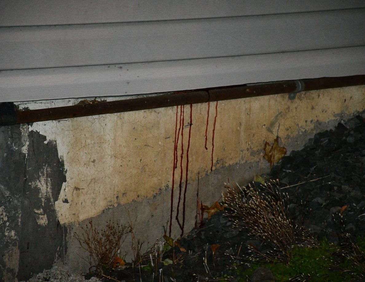 Dark reddish vertical streaks on a concrete foundation beneath a horizontal pipe and gray siding.