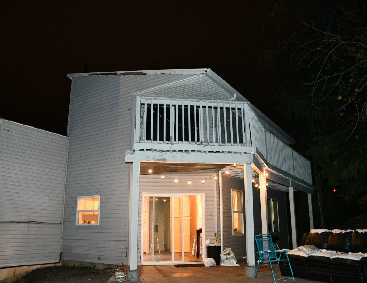 Gray house exterior at night with raised deck, lit sliding doorway and patio furniture.