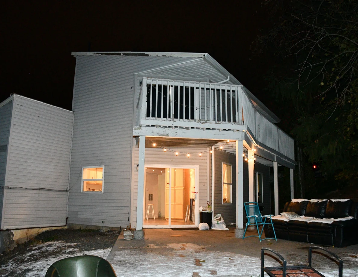 Wider night view of a gray house, elevated deck, lit patio doorway and outdoor sofa.
