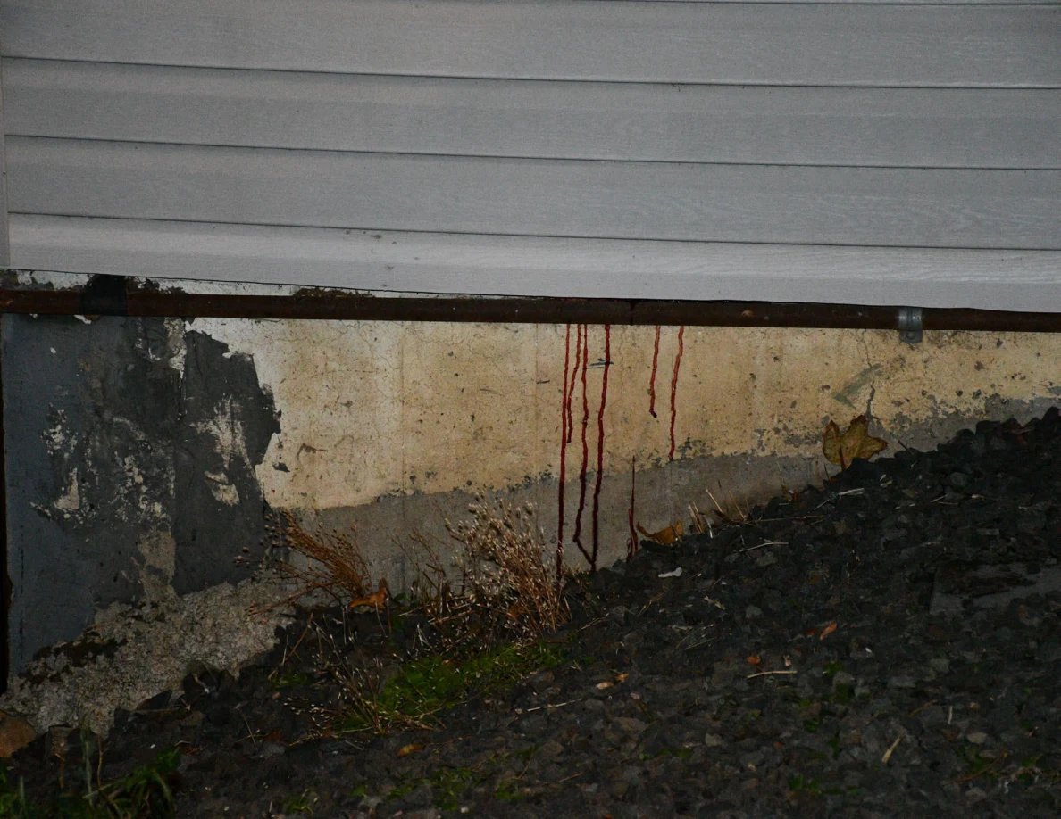 Concrete foundation with dark reddish streaks beneath a pipe, beside gravel and plants.