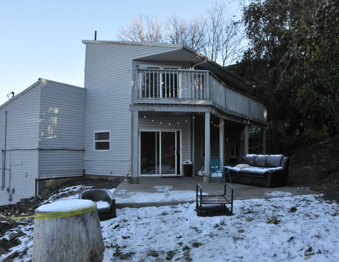 Wider daylight view of gray house, raised deck, snow-covered ground and patio seating.