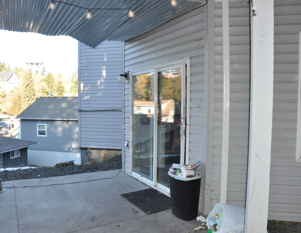Covered patio with string lights, sliding glass door and black trash bin.