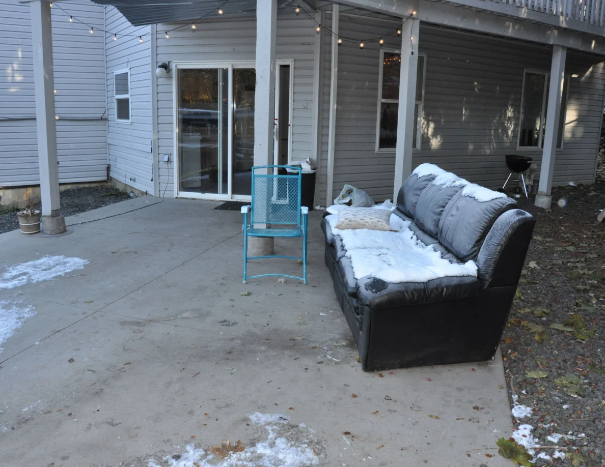 Snow-covered black sofa and turquoise chair on a concrete patio beneath string lights.