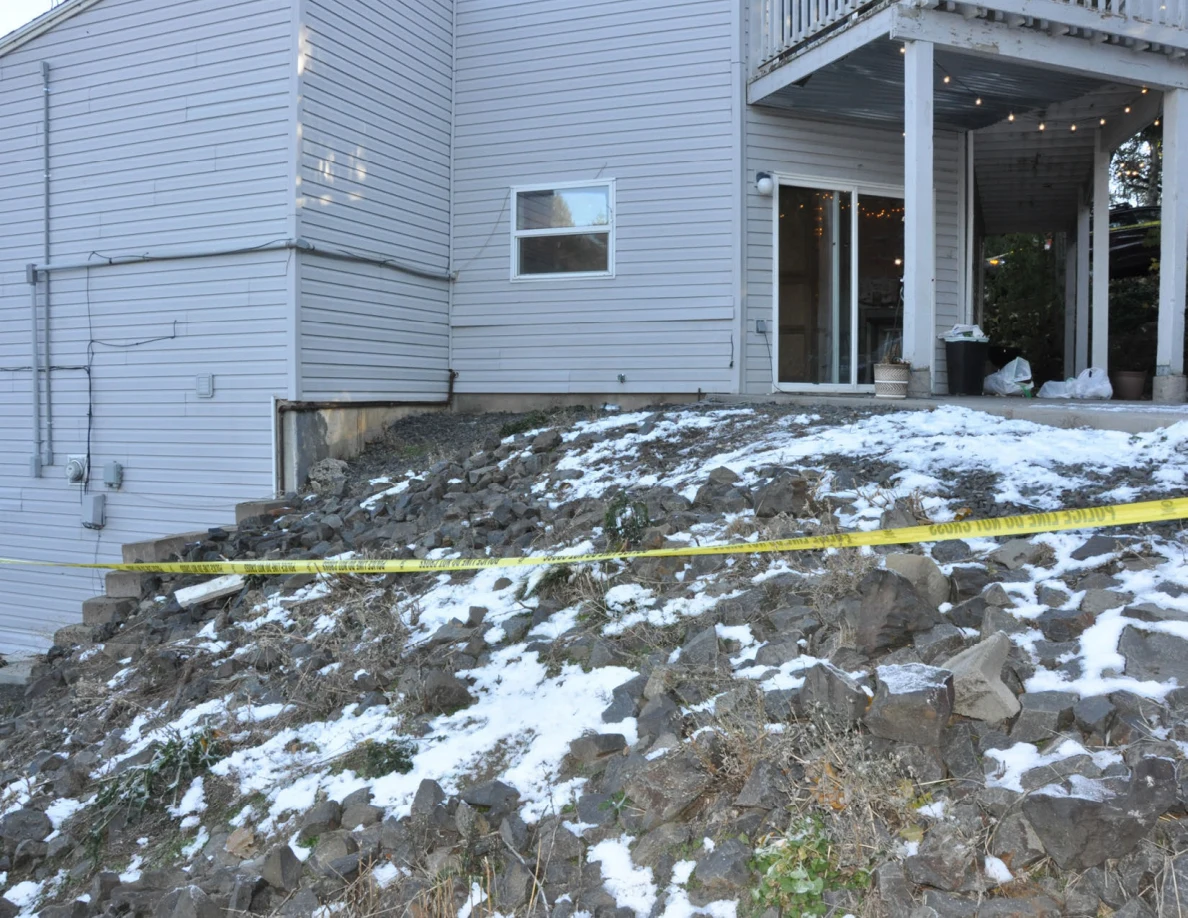Gray house above a rocky slope with patches of snow and yellow police tape.