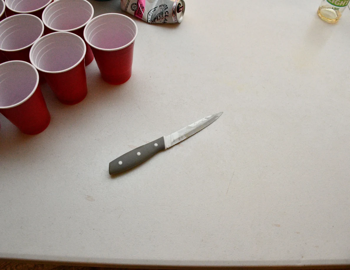 Small serrated kitchen knife with dark handle on a white table beside red plastic cups and beverage cans.