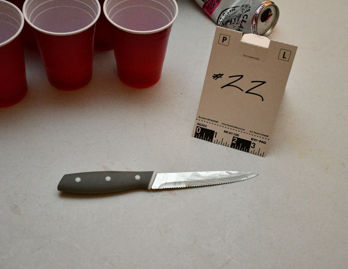 Dark-handled serrated knife on a white table beside card 22, red cups and a drink can.