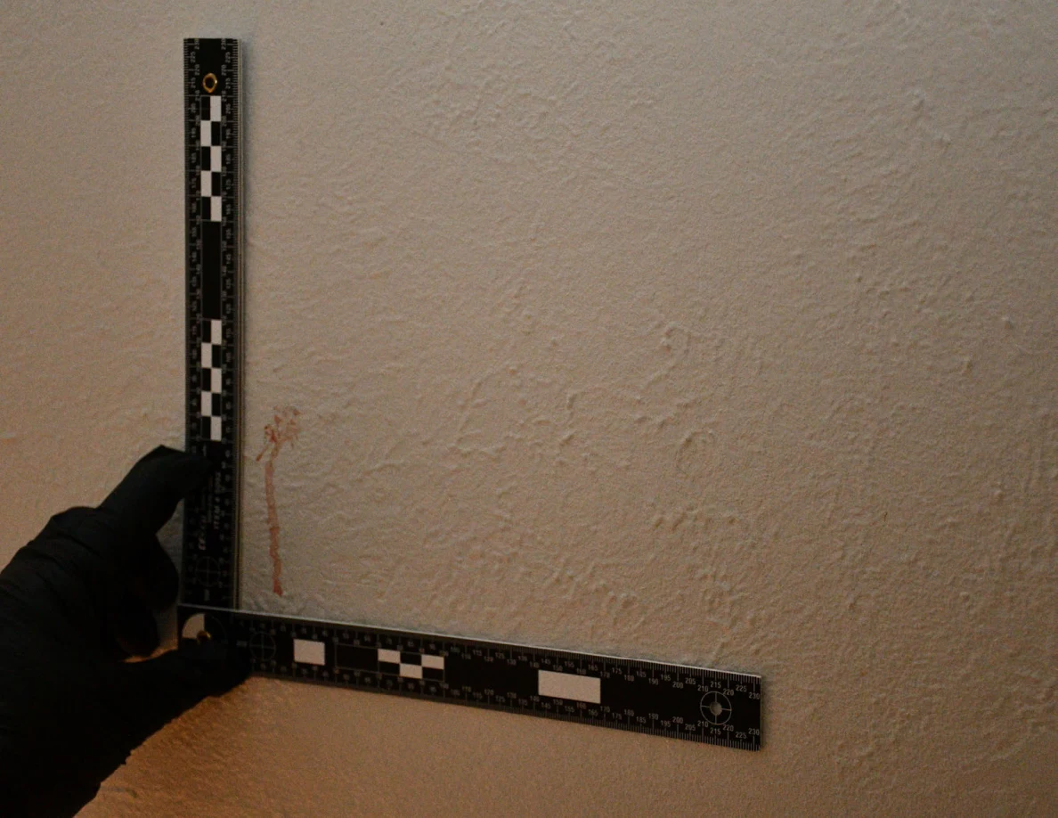L-shaped measurement ruler held beside a reddish vertical mark on a textured wall.
