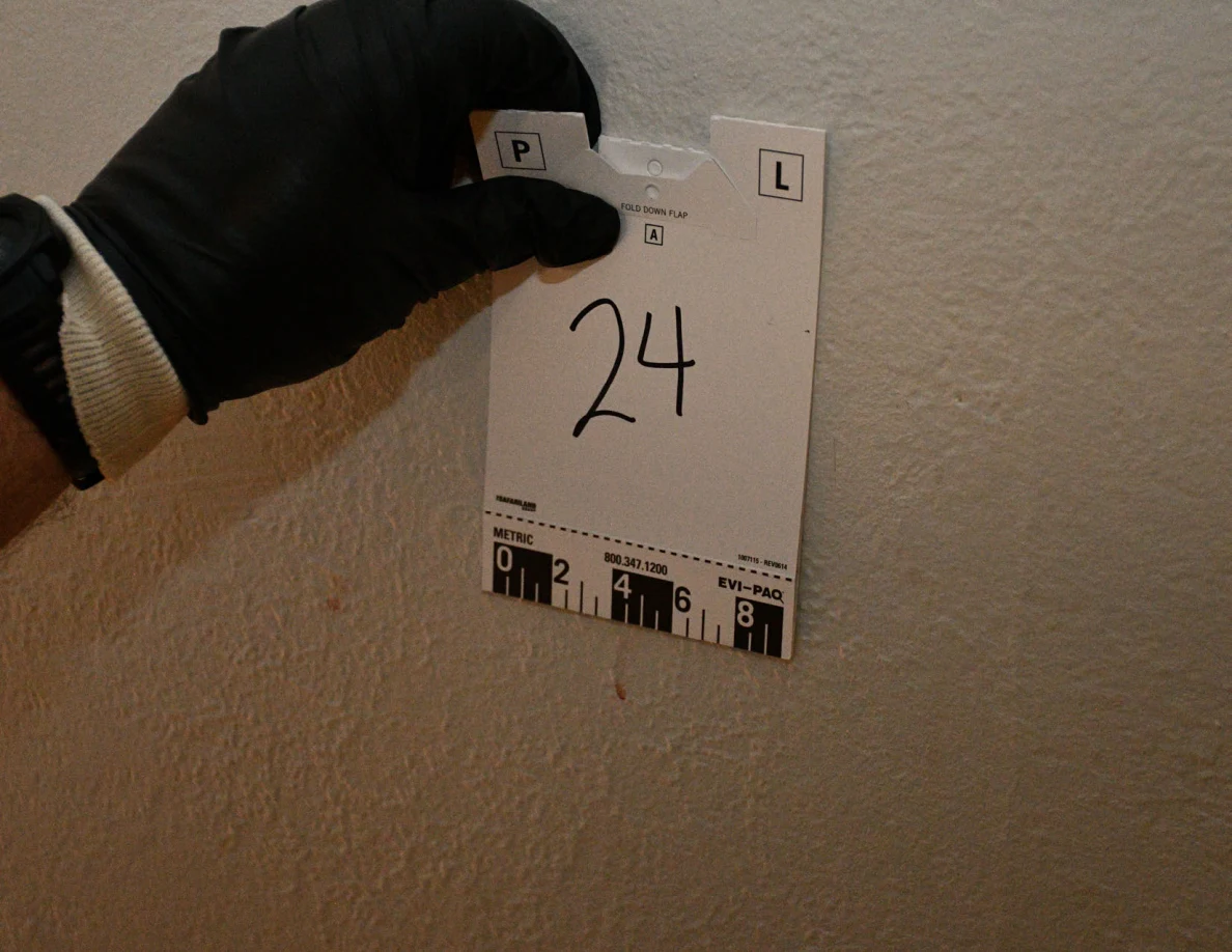 Black-gloved hand holding card 24 against a pale wall above a small reddish mark.