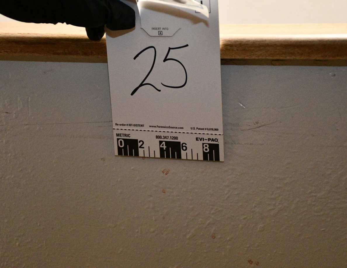Black-gloved hand holding card 25 beneath a wooden half-wall cap, beside small reddish spots.