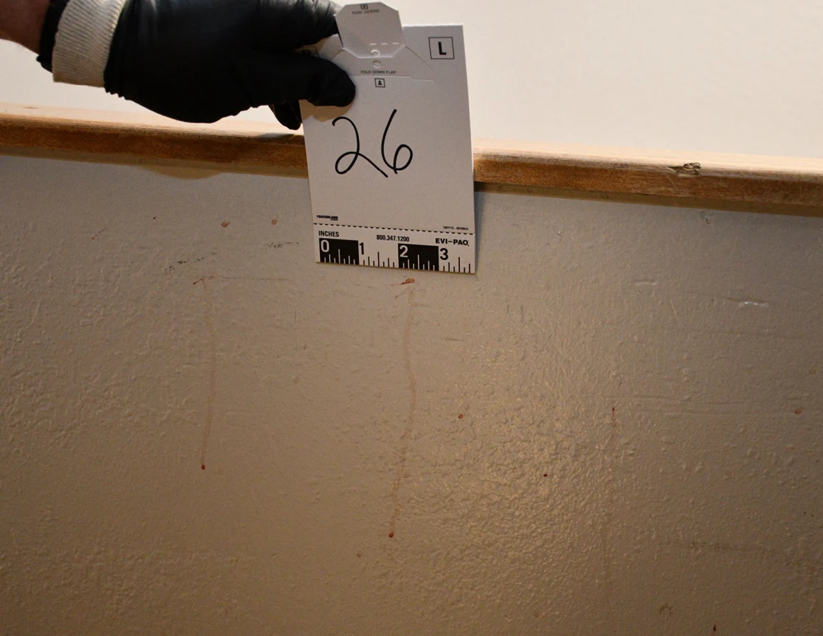 Black-gloved hand holding card 26 above thin reddish streaks on a half-wall.