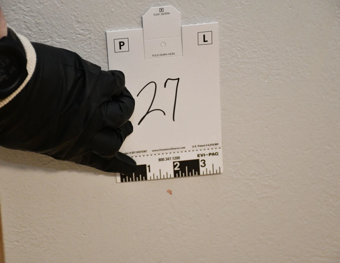Black-gloved hand holding card 27 beside a small reddish mark on a pale wall.