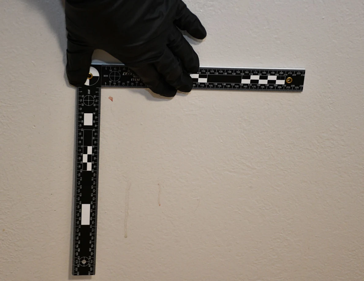 L-shaped ruler held beside a faint reddish mark on a pale textured wall.