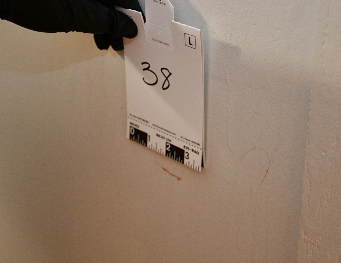 Black-gloved hand holding card 38 beside a reddish wall streak.