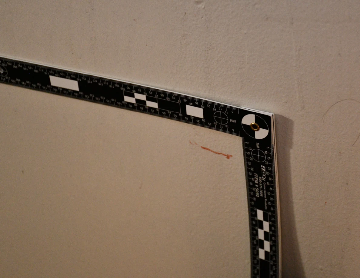 L-shaped measurement ruler beside a short reddish streak on a pale wall.