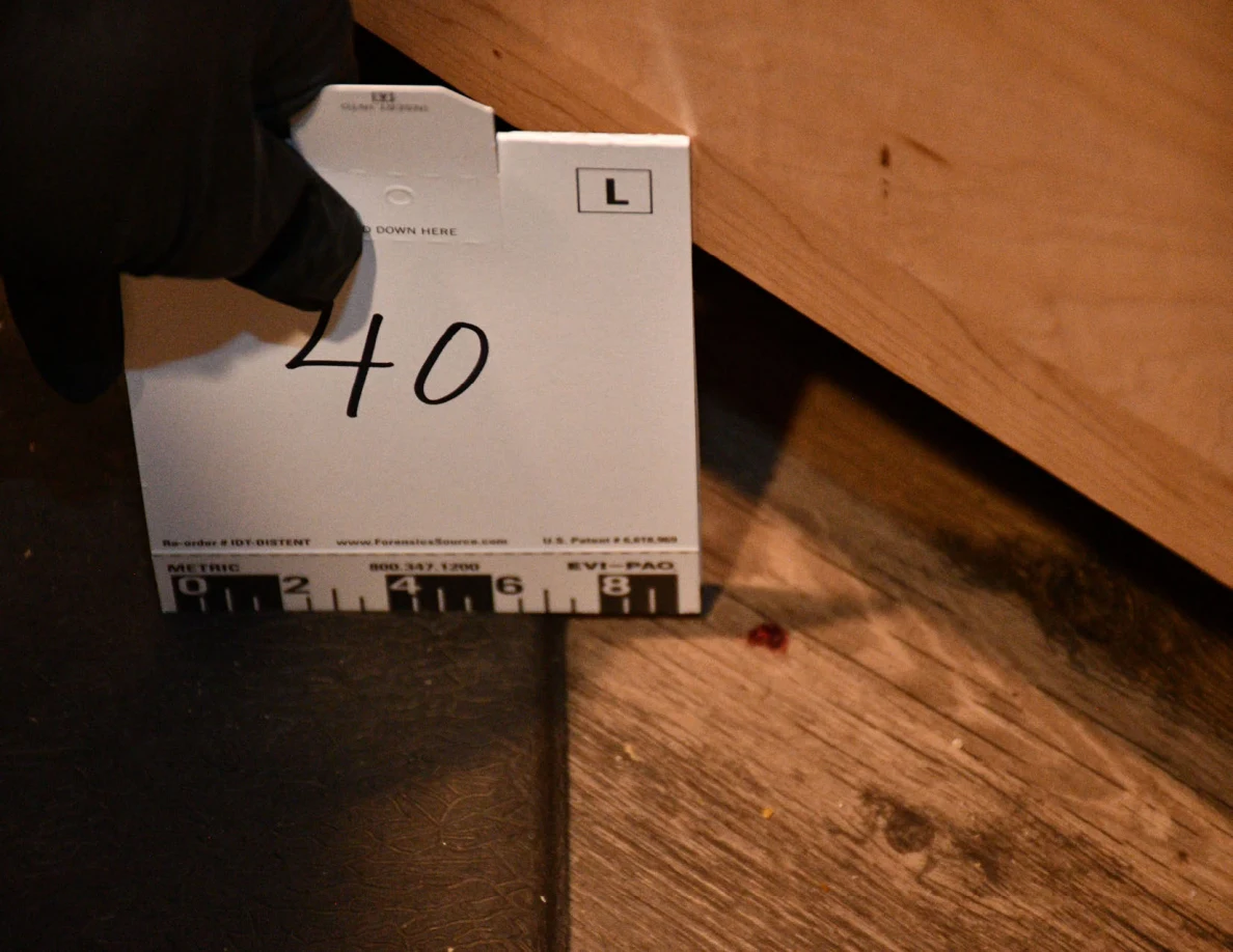 Black-gloved hand holding card 40 beside a small reddish spot on kitchen flooring.