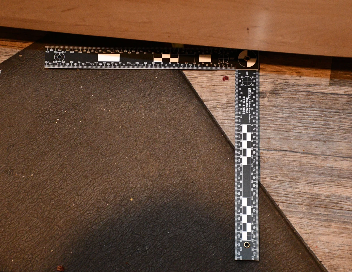 L-shaped ruler beside a small reddish floor spot at the edge of a dark kitchen mat.