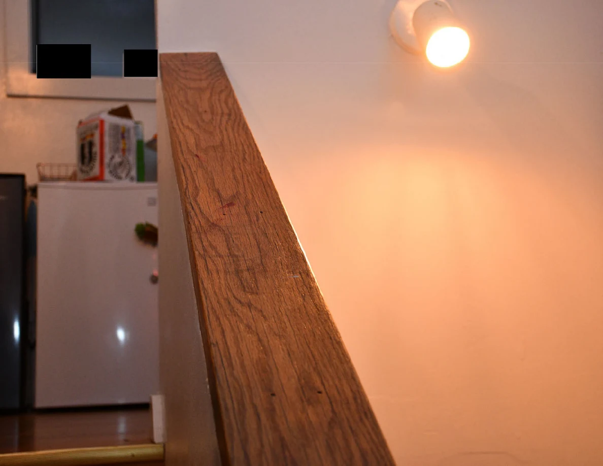 Wooden stair cap beneath a wall light, with refrigerator visible beyond and small black redactions.