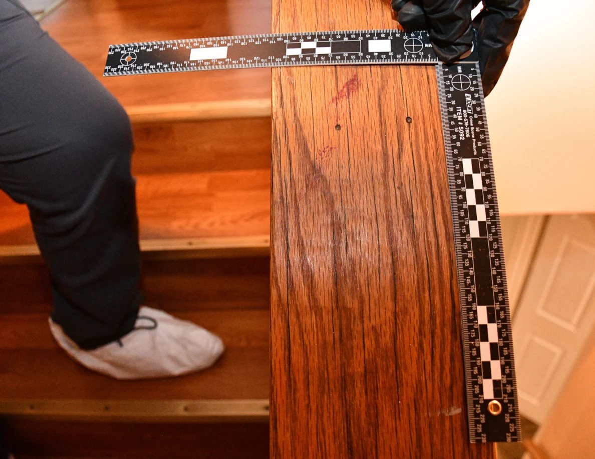 L-shaped ruler held beside reddish marks on a wooden stair cap, with stairs below.