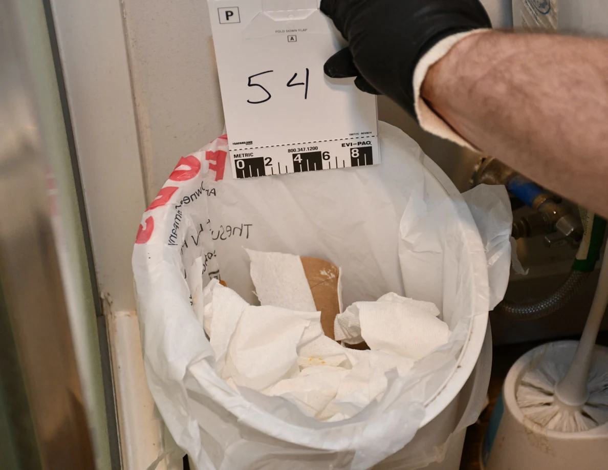 Card 54 held above a lined bathroom trash bin containing tissues and a cardboard roll.