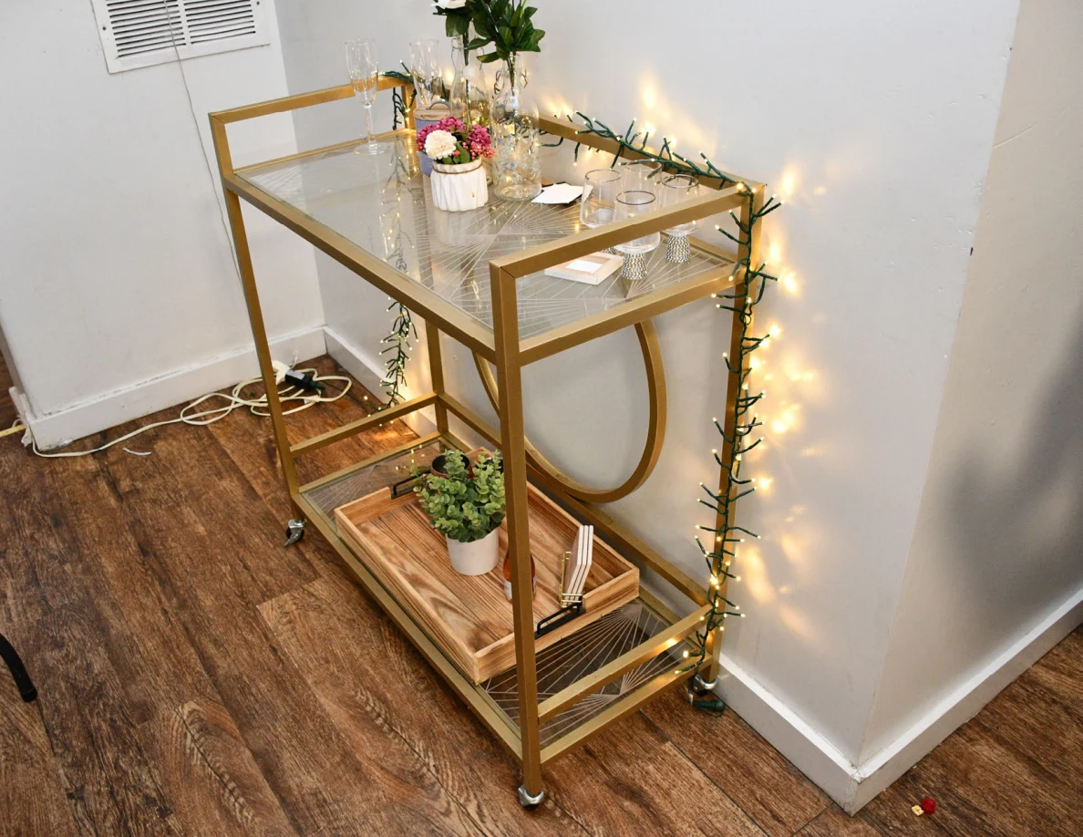 Gold-colored glass serving cart with plants, glassware and decorative string lights.