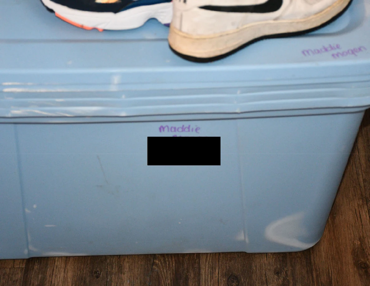 Close view of a blue storage tote with handwritten label partly blacked out and shoes on top.